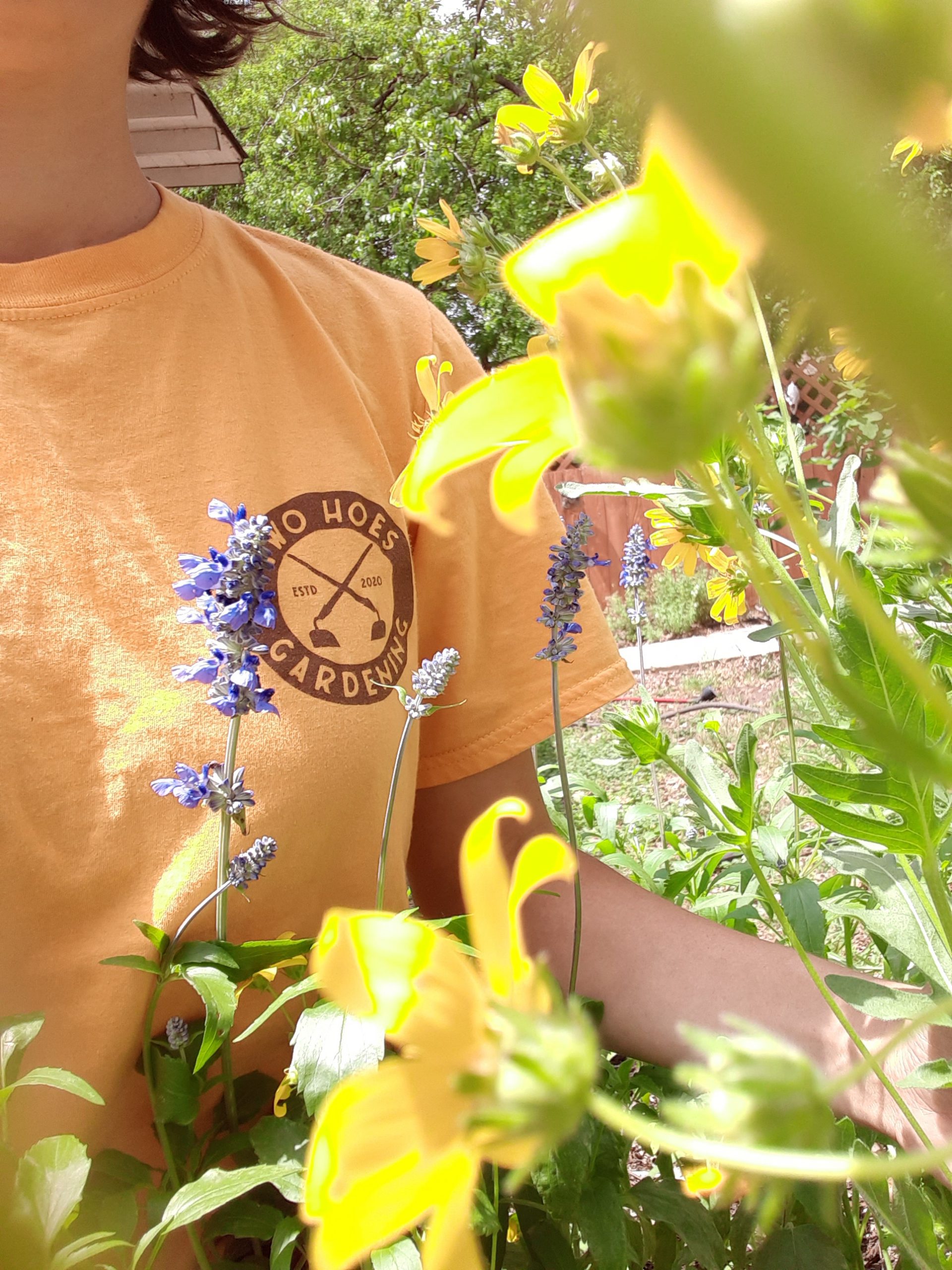 in frame, woman's torso wearing a yellow shirt with brown two hoes gardening logo positioned right behind lush wildflowers